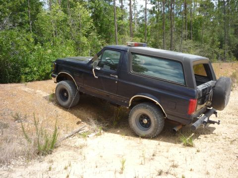 1990 Ford Bronco - This dark blue SUV, likely a 1992 model, is parked on a dirt road surrounded by trees.