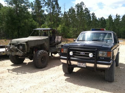 1990 Ford Bronco - Two off-road vehicles, one a black Ford Bronco, are parked on a dirt lot with trees in the background.