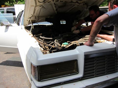 1991 Ford LTD Crown Victoria - Two men are attending to the engine bay of a white sedan, with another vehicle partially visible in the background.