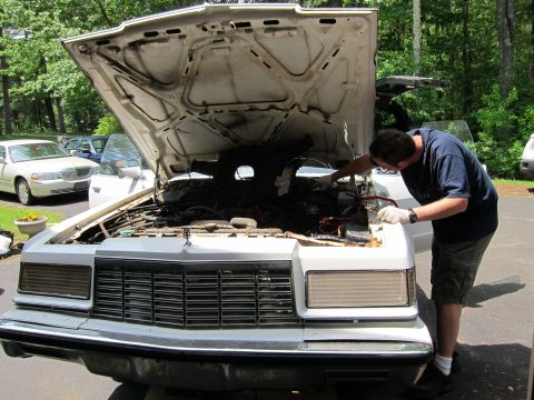 1991 Ford LTD Crown Victoria - Man works on the engine of a white sedan, with another car visible in the background.