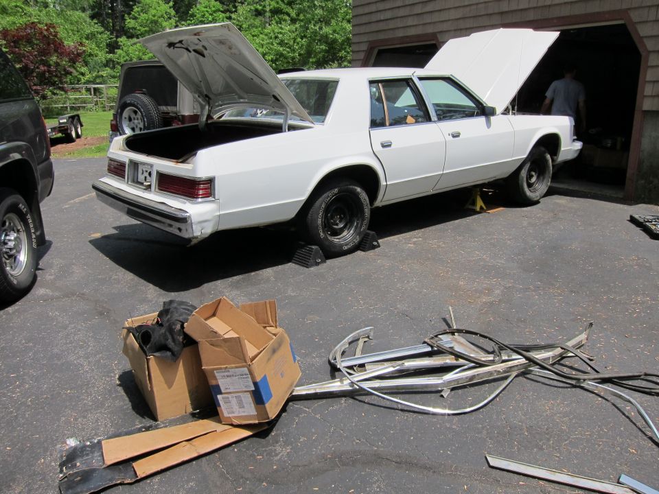 White sedan with hood and trunk open, parked in a driveway next to a garage. Parts are laid out on the ground