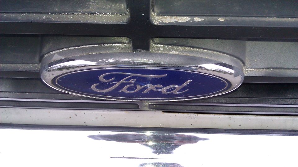 Ford emblem on a chrome grille with black horizontal bars. The emblem is blue with silver script