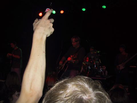 1989 Ford Bronco - A band plays a live music set on stage, with a hand in the foreground holding up a bottle cap.