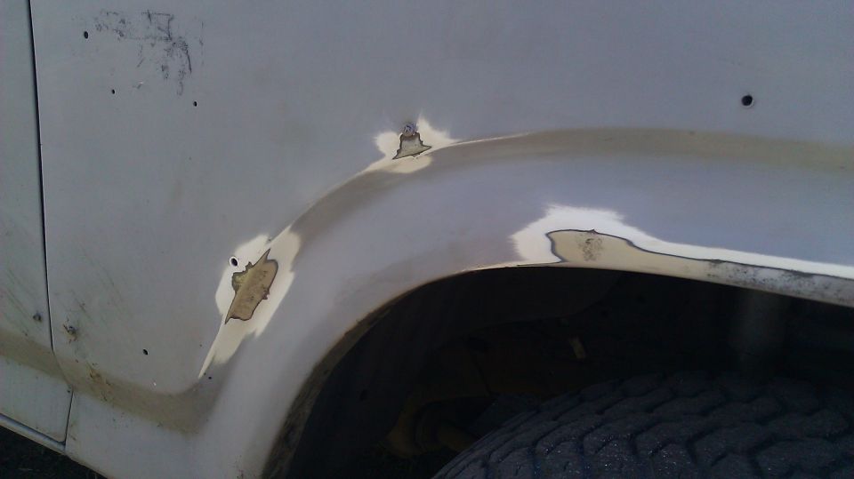 White vehicle fender with rust patches and primer, showing damage around the wheel well