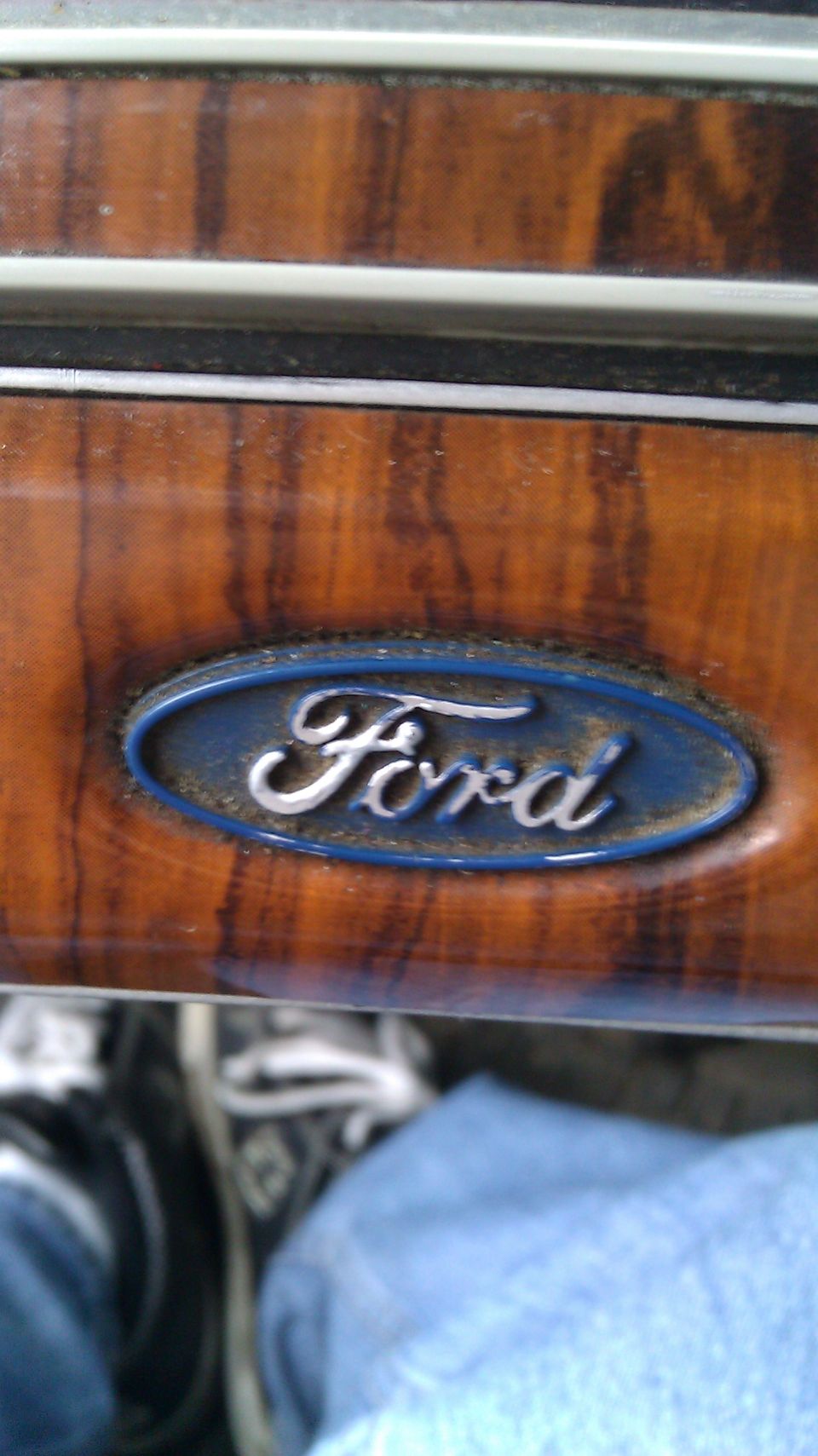 Ford oval logo on wood grain dashboard trim. Jeans and shoes visible below