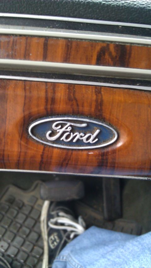 1984 Ford Bronco - Close-up of a Ford emblem on a dashboard with wood-grain trim. A foot is visible on the pedal.