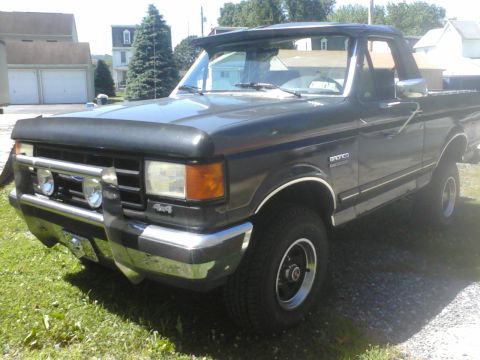 1988 Ford Bronco - A dark gray Ford Bronco is parked on a grassy area with a house and trees in the background.