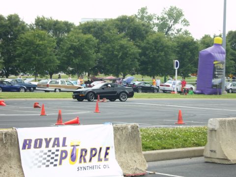 2007 Ford Mustang - A black 2007 Ford Mustang navigates a course marked by orange cones, with other vehicles and a large inflatable bottle in the background.