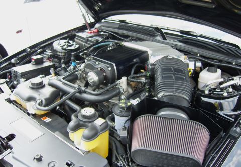 2007 Ford Mustang - The engine bay of a black 2007 Ford Mustang is visible, a prominent supercharger and aftermarket air intake system.
