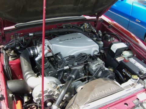 2007 Ford Mustang - The engine bay of a red car, a prominent supercharger and intake system. Another vehicle is partially visible in the background.