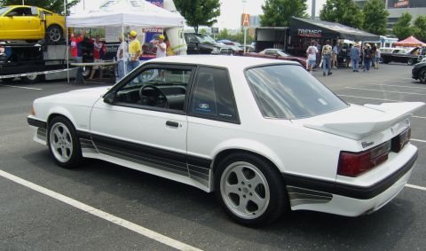 2007 Ford Mustang - A white Ford Mustang from the late 1980s is parked at an outdoor event with other vehicles and tents.