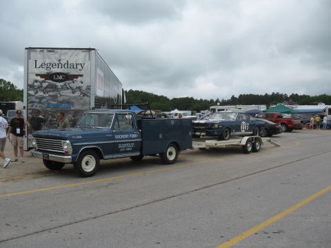 1968 Shelby GT500 - A blue Ford pickup truck tows a blue Ford Mustang race car on a trailer at an outdoor event.