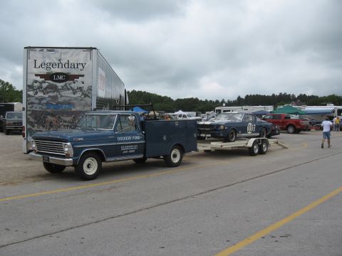 1968 Shelby GT500 - A blue Ford truck tows a trailer carrying a blue 1960s muscle car, possibly a Mustang, with racing stripes and the number 03 visible.