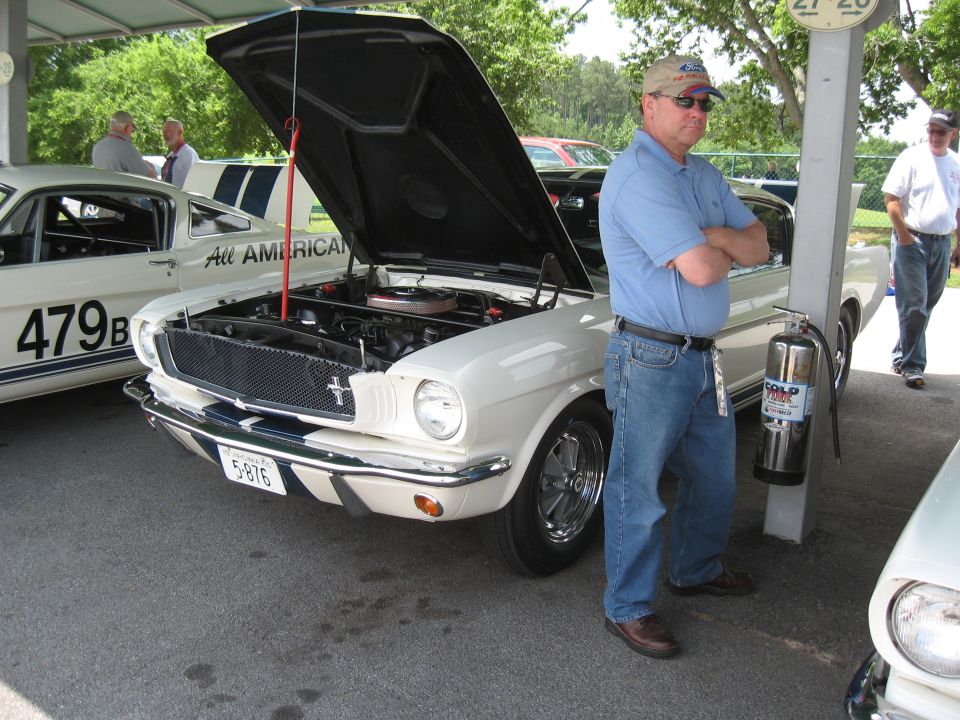 Guy in blue shirt and jeans stands by a white Ford Mustang with its hood open. Another Mustang with 'All American' and '479B' is to the left