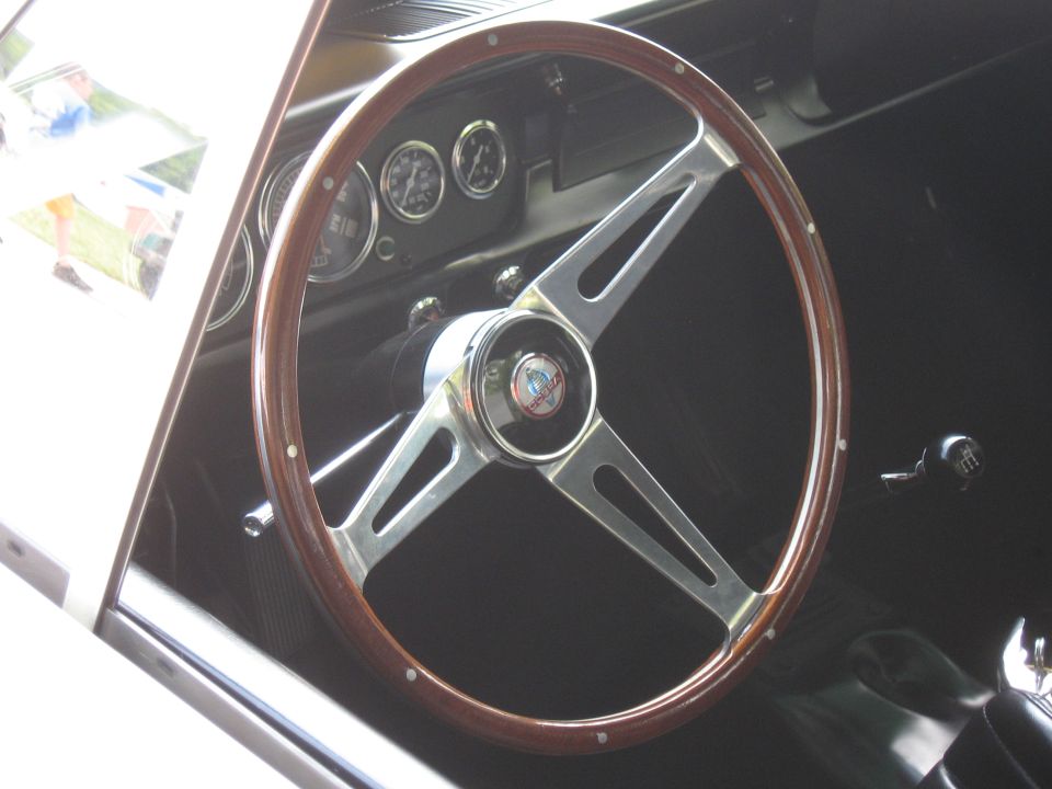 Classic car interior showing a wood-rimmed steering wheel with a chrome horn button and three gauges on the dashboard