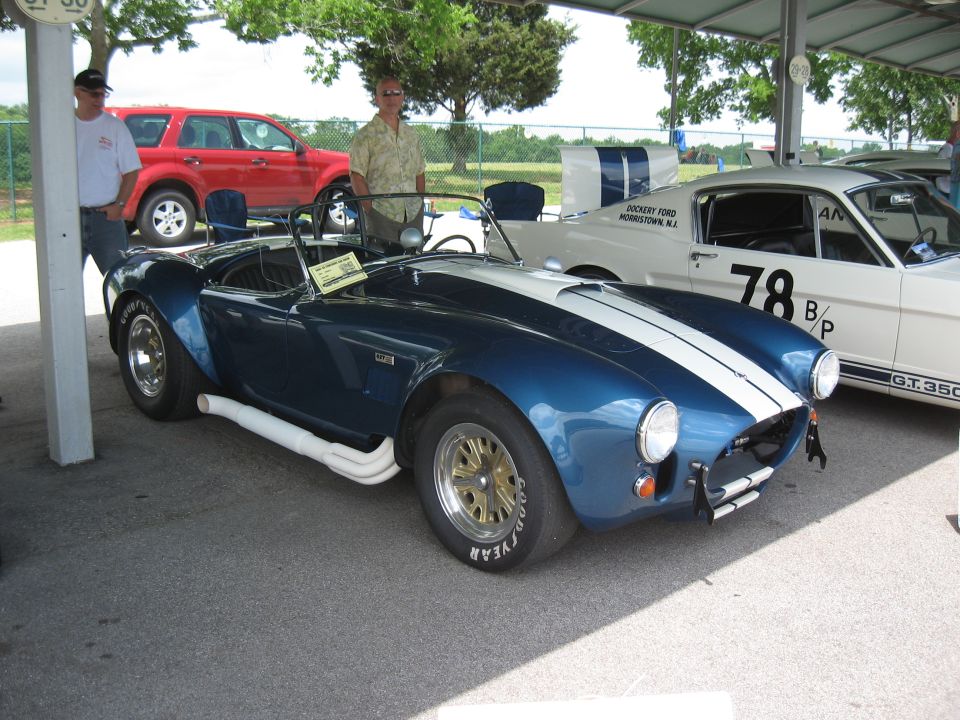 Blue convertible sports car with white stripes, side pipes, and gold wheels parked under a canopy