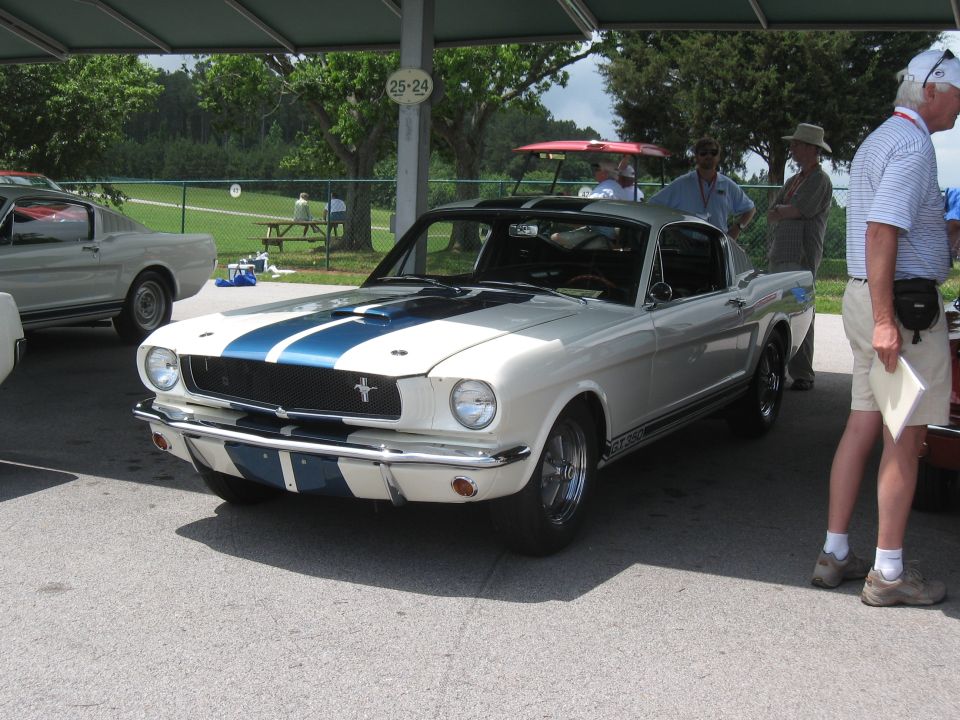 White 1965 Shelby GT350 with blue stripes parked under a canopy. Another classic car is visible to the left