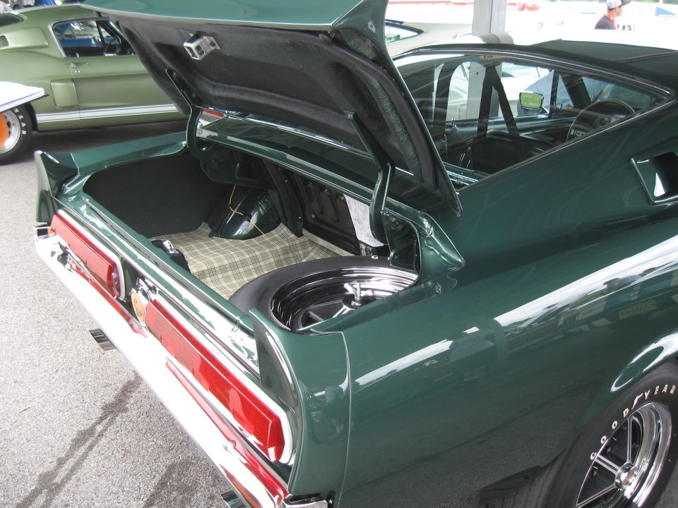 Green 1968 Shelby GT500 with trunk open, showing spare tire and plaid liner. Another green Shelby is visible in the background