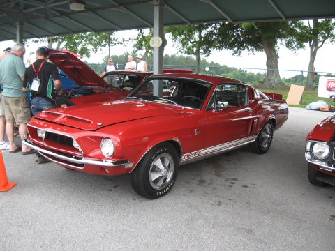 1968 Shelby GT500 - A red 1968 Shelby GT500 Mustang is displayed at an outdoor event, with other vehicles and attendees present.