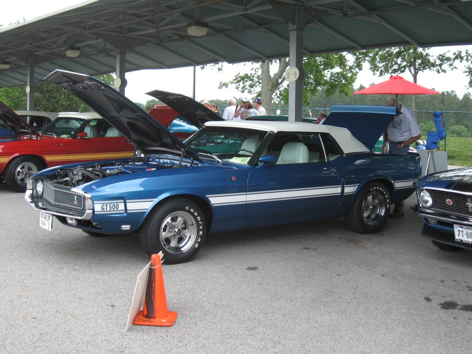 Blue 1969 Mustang convertible with white stripes and a white top. Hood and trunk are open. Another car is parked to the right