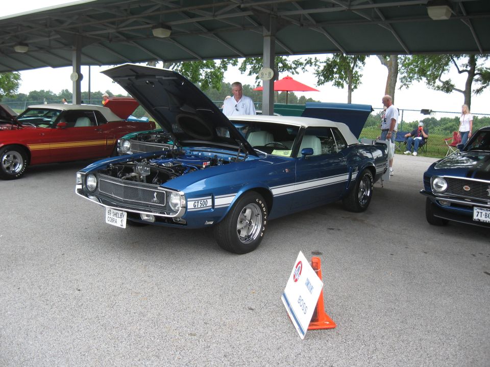 Blue 1969 Shelby GT500 convertible with white stripes, hood open, parked under a canopy