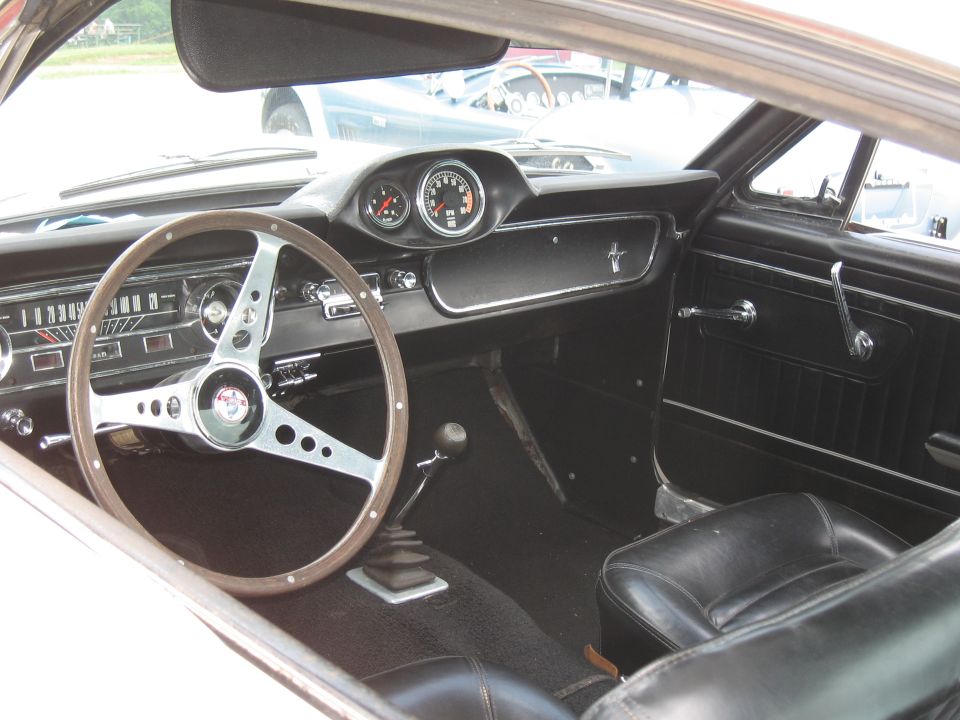 Interior of a classic Mustang, showing the dashboard, steering wheel, and seats. The dashboard has a tachometer and speedometer