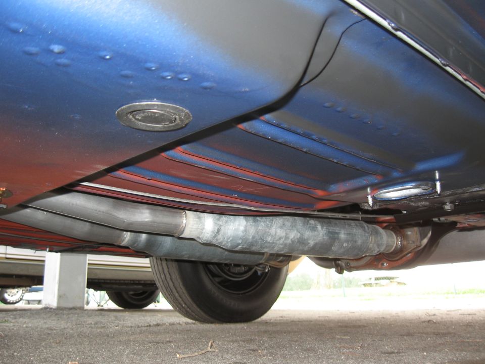 Underside of a car showing the exhaust pipes and part of a wheel. There's a SuperMotors logo at the bottom right