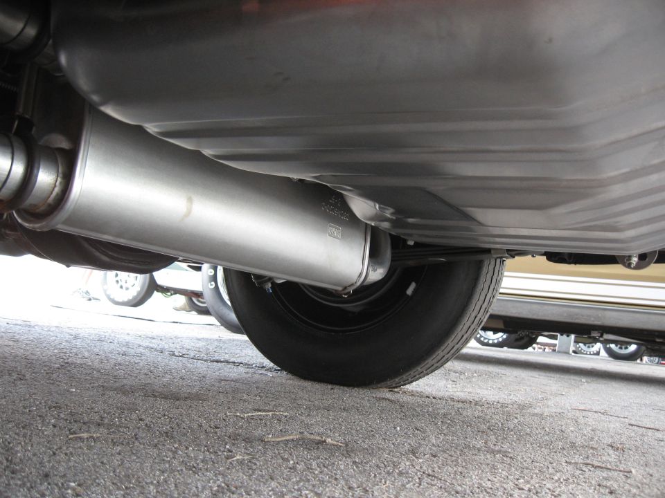 Underside view of a car's exhaust pipe and muffler, with a tire visible. Another car is in the background