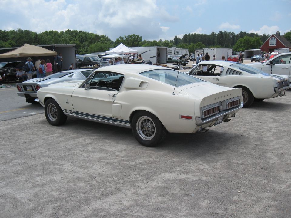 Two white 1968 Shelby GT500s parked on gravel lot with tents and trailers in background