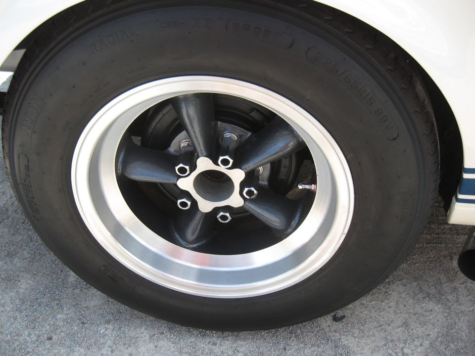 Close-up of a black five-spoke wheel with a polished lip and a radial tire on a white car with blue stripe