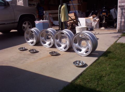 1988 Ford Bronco - Four silver alloy wheels and three hubcaps are laid out on a concrete driveway next to a parked truck.