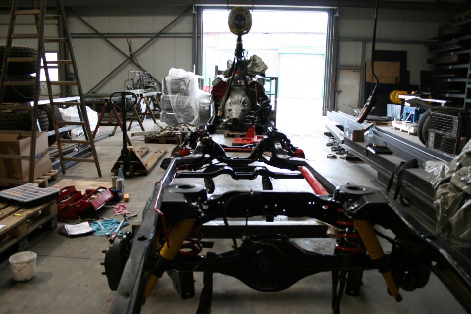 Vehicle chassis and engine suspended in a workshop, with tools and tires scattered around