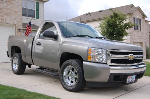 2007 Chevrolet Silverado 1500 - A silver pickup truck is parked in a driveway with a house in the background. An American flag is visible in the truck bed.
