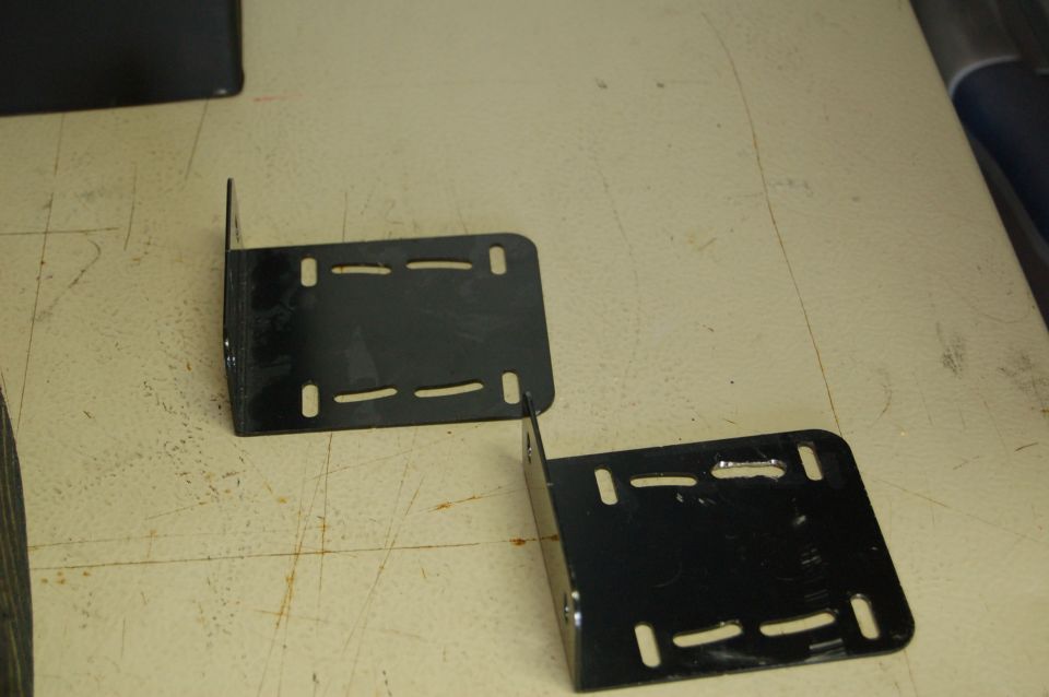 Two black metal L-brackets with slotted holes on a textured surface