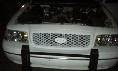 1998 Ford Crown Victoria - The front of a white 1998 Ford Crown Victoria is visible with its hood open, revealing the engine compartment and a distinctive grille.