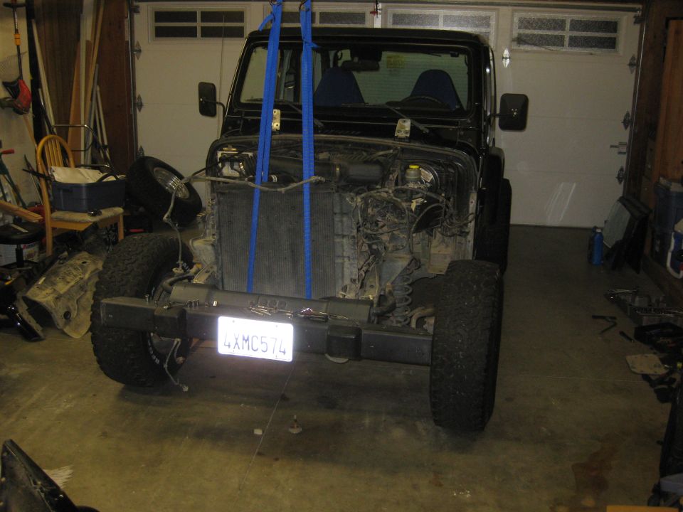 Black Jeep Wrangler with front end disassembled, suspended by blue straps, showing engine components and a license plate reading 4XMC574
