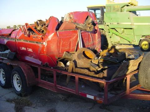 1992 Ford Bronco - A severely damaged red pickup truck cab, labeled 'LAWN CARE', is loaded onto a trailer beside a large green combine harvester.