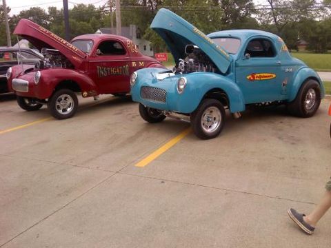 1970 Ford Ranchero - Two vintage hot rod coupes, a red 'Instigator' and a blue 'Schumacher's', parked side-by-side with hoods open.