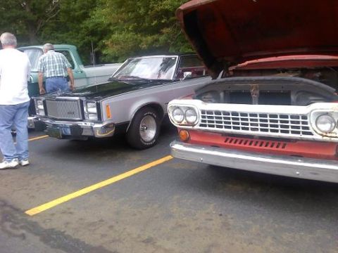 1970 Ford Ranchero - A silver and black coupe with a prominent chrome grille is parked next to a red truck with its hood open.