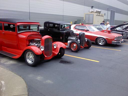 Several hot rod cars are parked in a lot, including a red sedan and a red and white coupe. A "SUPERMOTORS" logo is visible