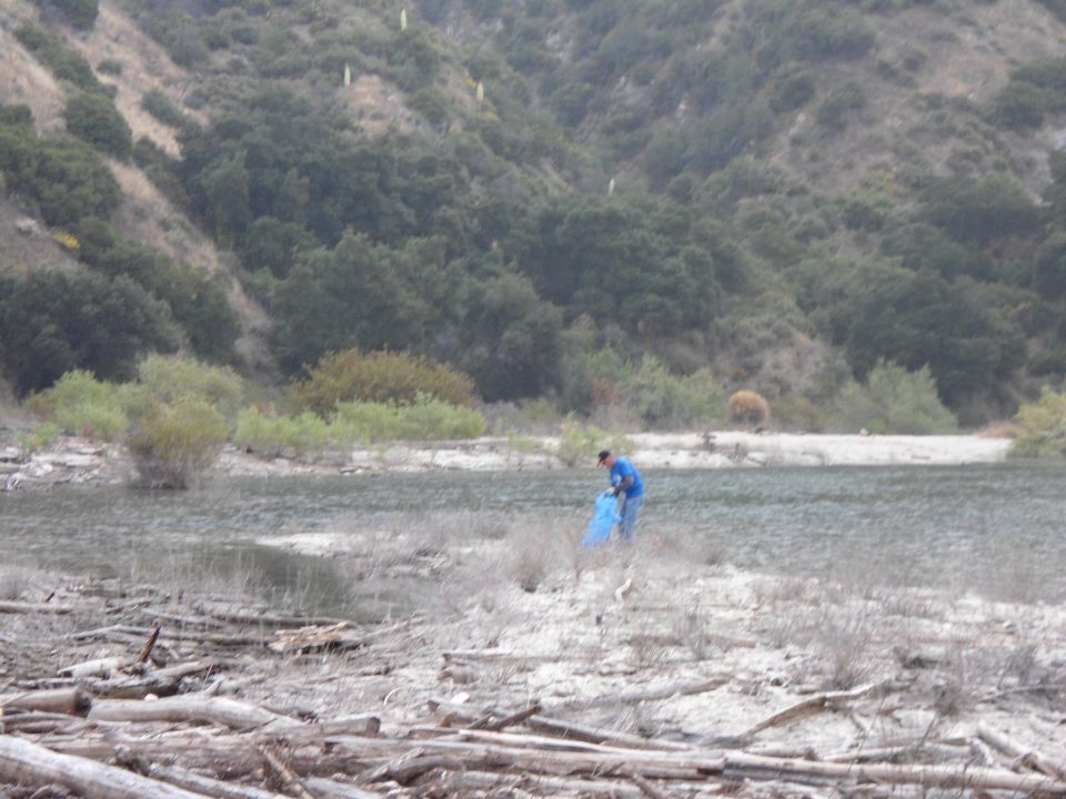 Person in blue jacket collecting trash with a blue bag near a lake, wooded hill behind
