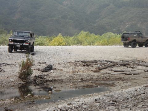 1994 Ford Bronco - Two SUVs are parked in a rocky, dry riverbed with a wooded hillside in the background.