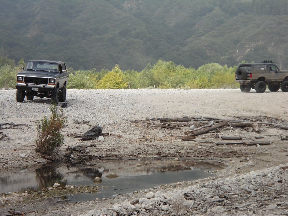 Black 1980s Ford Bronco parked on gravel, with a six-wheeled SUV in the background