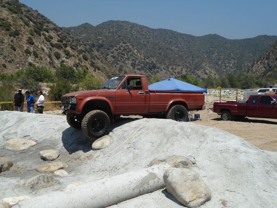 Brown Toyota pickup truck on rocky terrain with a blue tarp in the bed. Another red pickup is parked behind it