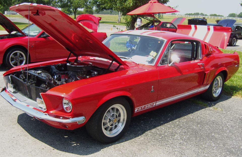 Red 1967 Mustang GT350 with hood open, white stripes on hood and trunk. Another red Mustang in background