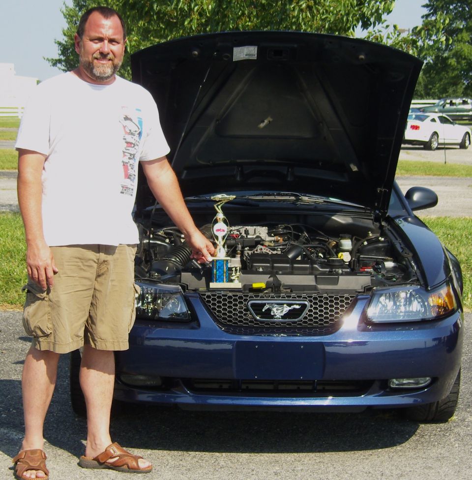 Man with trophy in front of blue Ford Mustang with hood open. Another white Mustang is parked in the background