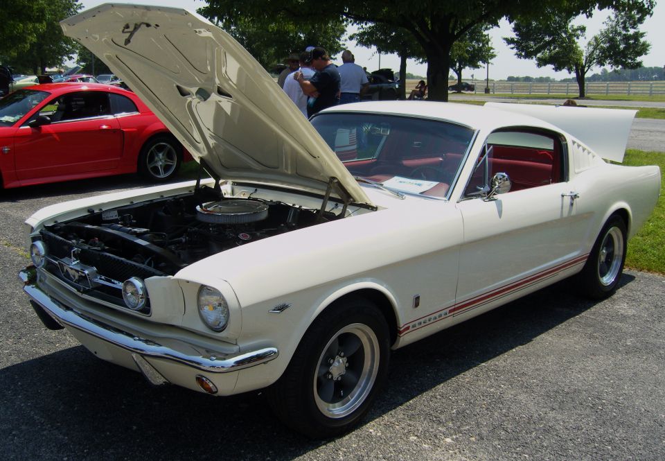 White classic Ford Mustang with red interior and red stripe, hood and trunk open. Red Ford Mustang visible in background