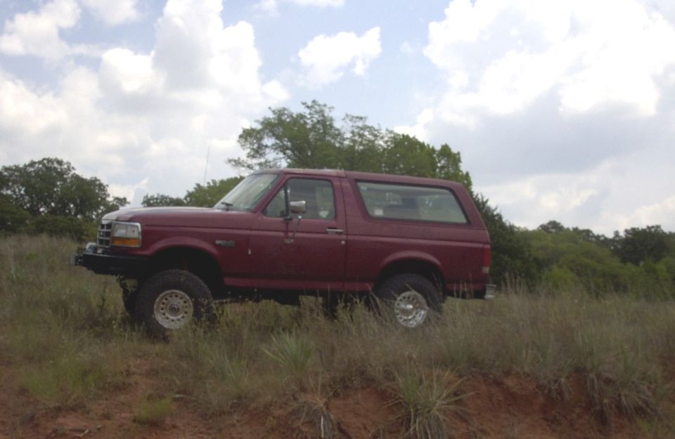 Maroon 1990s full-size Ford Bronco parked on a grassy hill under a cloudy sky