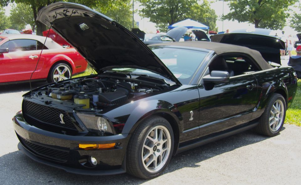 Black convertible Mustang with hood open, parked next to a red Mustang. Engine visible