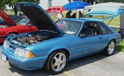 2007 Ford Mustang - A blue 1987-1993 Ford Mustang 5.0 is displayed with its hood open at an outdoor car show, alongside other vehicles.